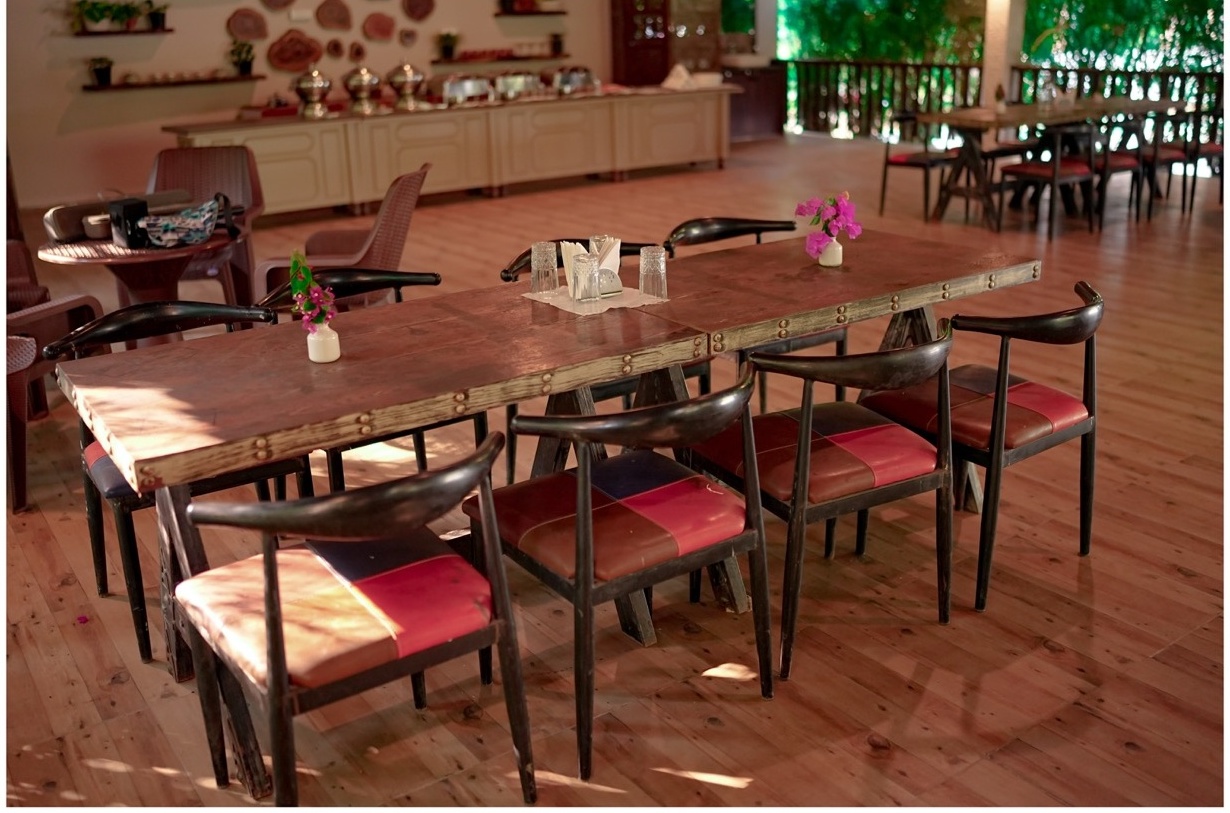 Rosewood Villas Pench wooden dining hall with long tables, chairs, and flower vases under warm lighting.
